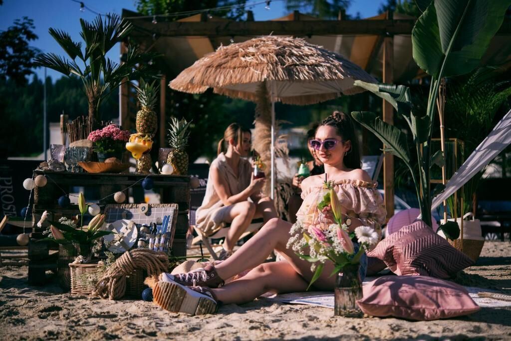 Escena de playa con una sombrilla de paja, plantas en macetas y un ambiente relajado.