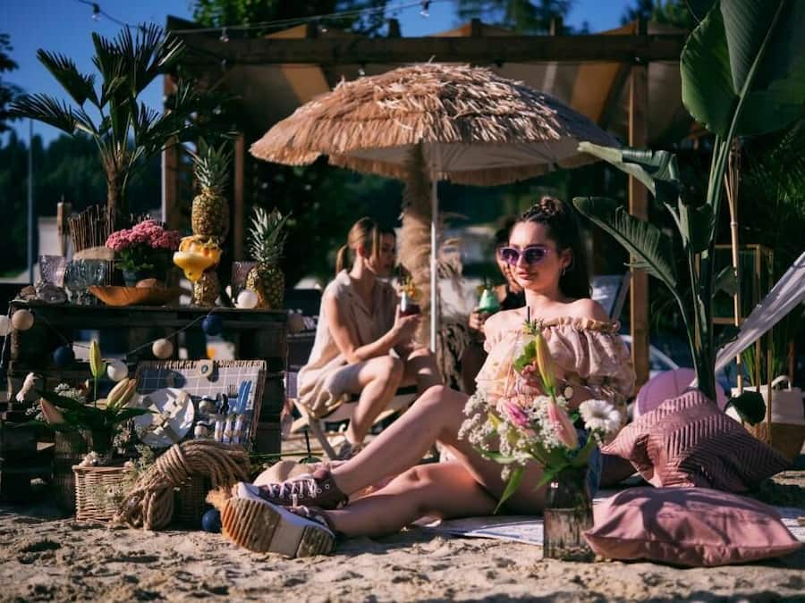 Escena de playa con una sombrilla de paja, plantas en macetas y un ambiente relajado.
