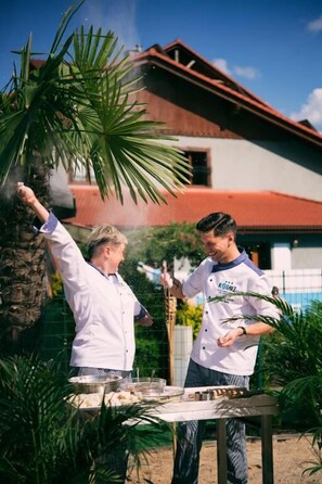 Two individuals outdoors near a table with cooking equipment, a palm tree, and a building with a red roof. - Hotel Kotarz Spa & Wellness (Brenna)