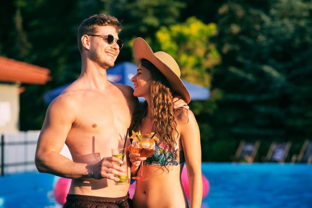 Una pareja junto a una piscina con bebidas, rodeada de vegetación.