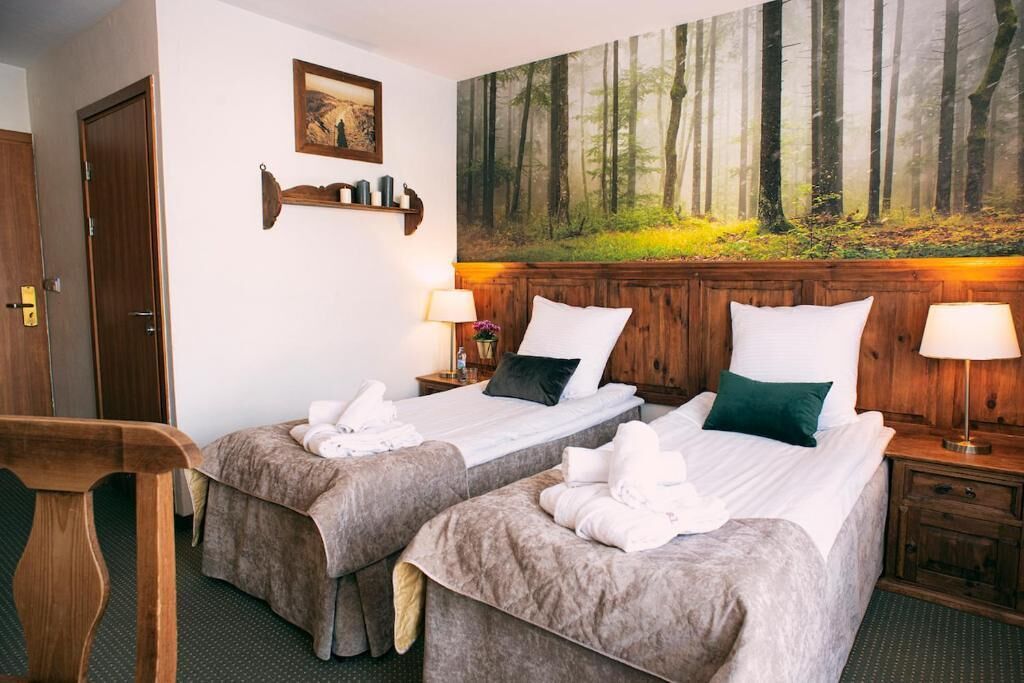 Habitación de hotel con dos camas, un cabecero de madera, un cuadro de un bosque y un pequeño escritorio con una silla.