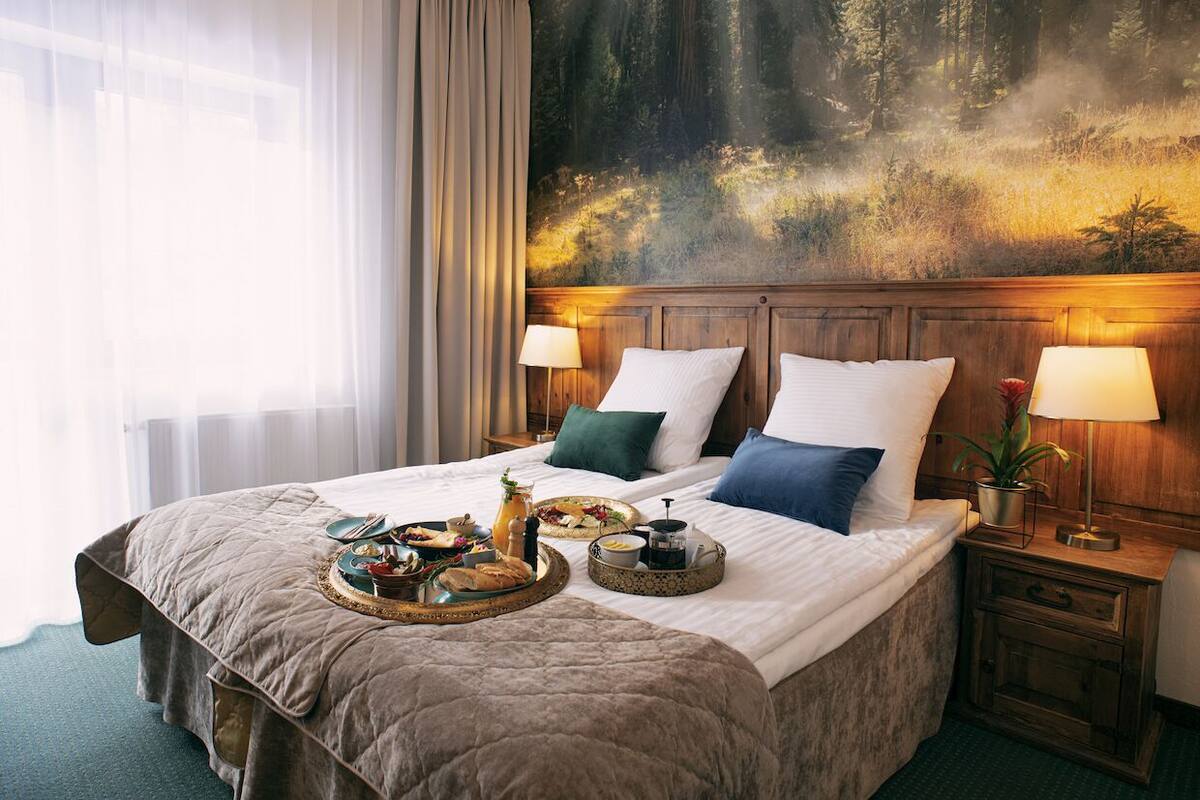 A hotel room with a large bed, two pillows, a nightstand with a lamp, and a painting of a forest on the wall.