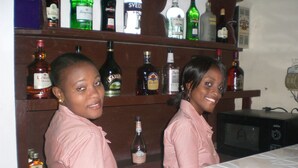 Two individuals standing behind a bar counter with various liquor bottles on shelves behind them. - Metro Residences Hotel (Cap-Haitien)