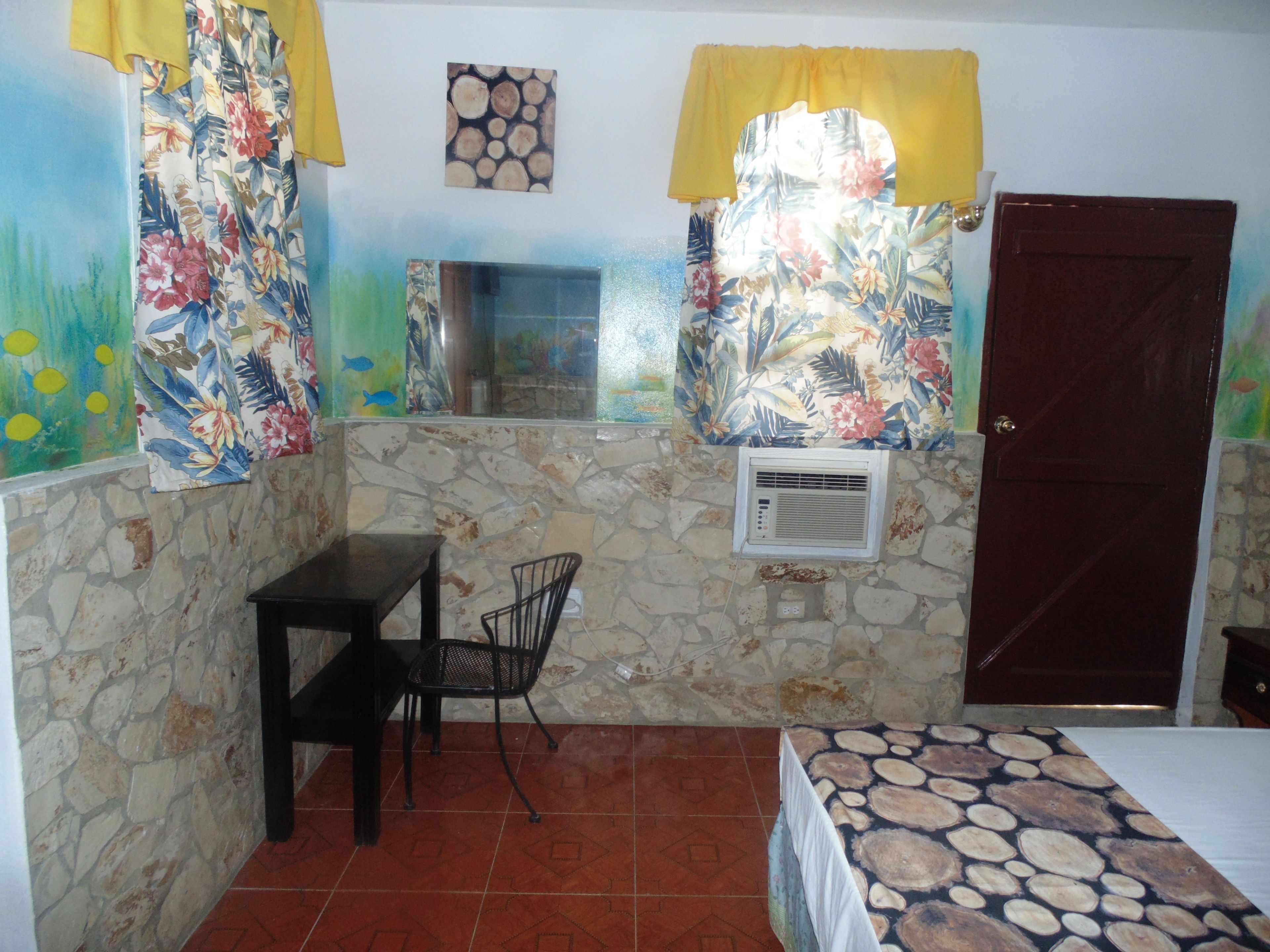 A room with a stone wall, a bed with a patterned bedspread, a small table with a chair, a window with a decorative curtain, and an air conditioning unit.