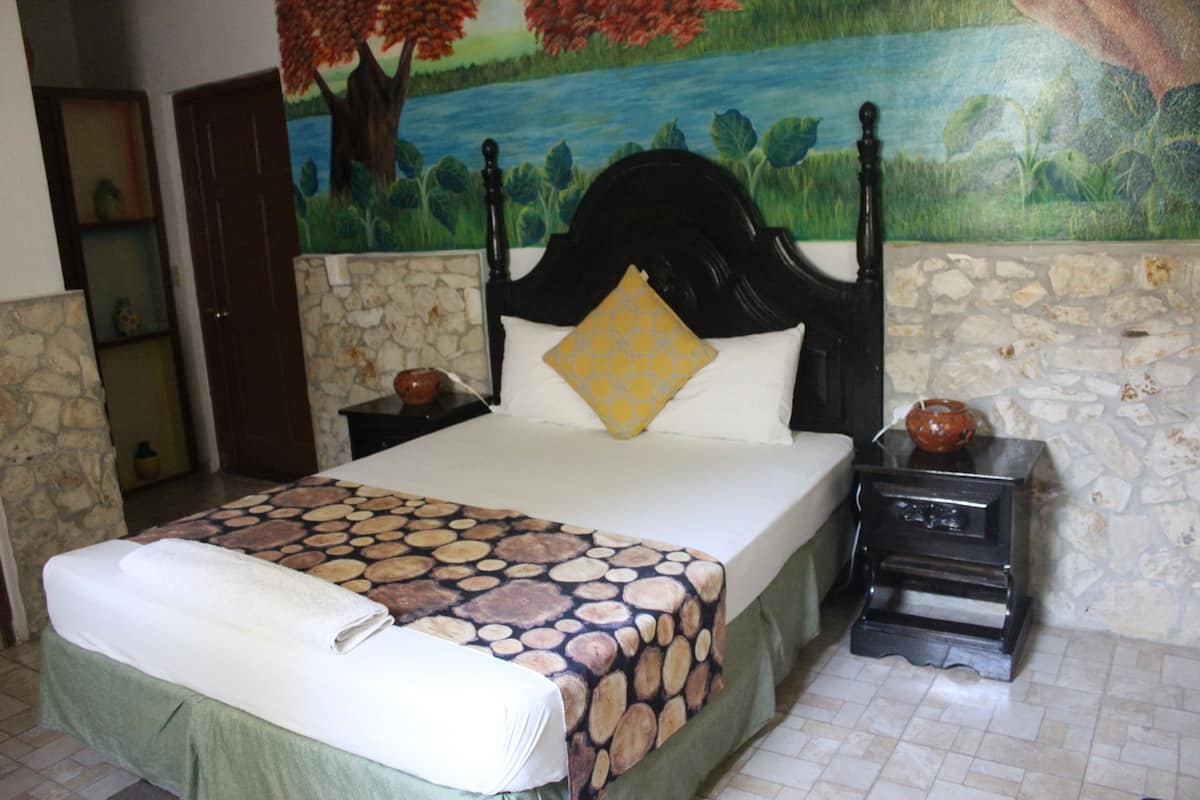 A bed with a patterned bedspread, a headboard with a decorative pillow, a nightstand with a vase, and a stone wall.