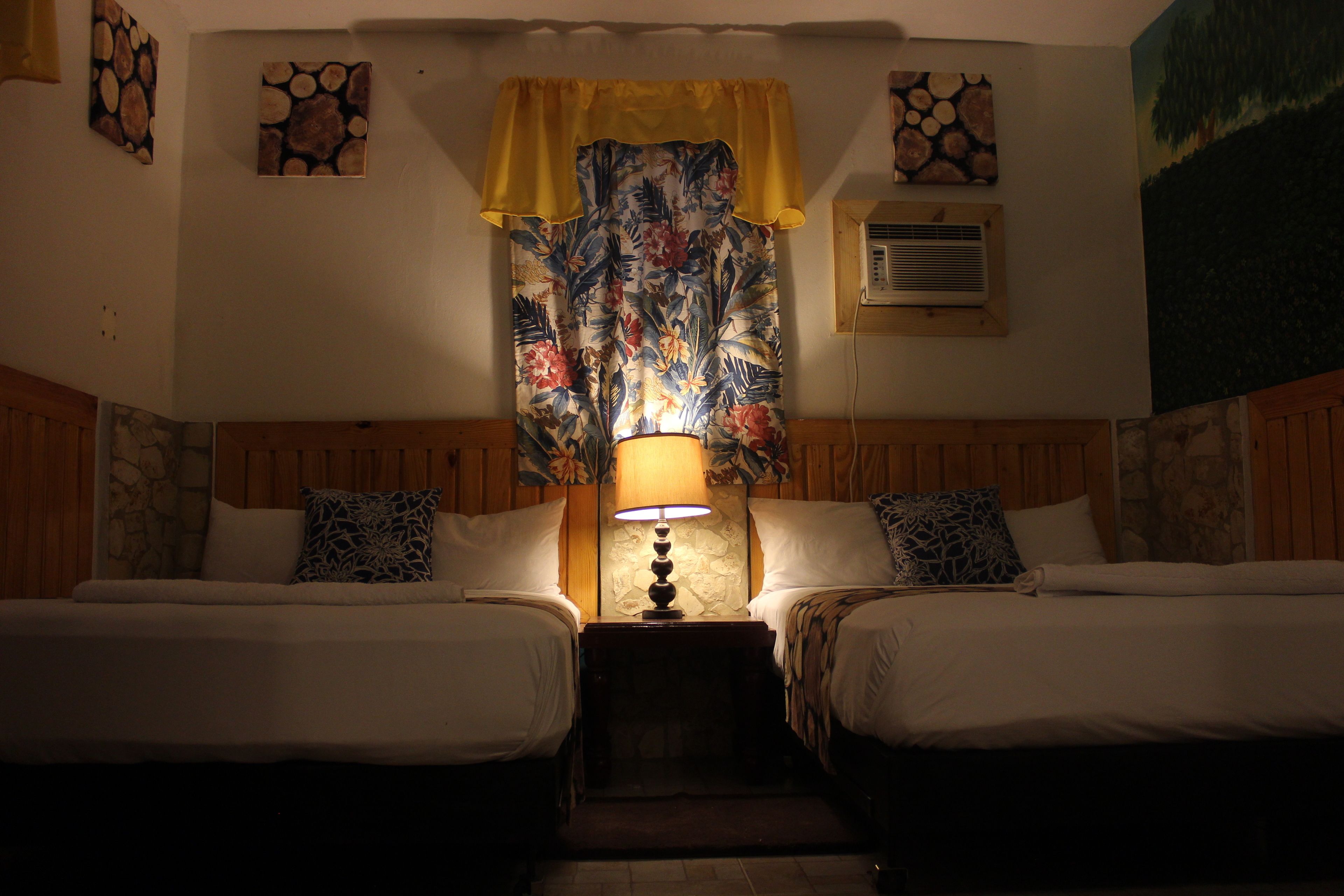 A room with two beds, a nightstand with a lamp, a window with a floral curtain, and artwork on the walls.