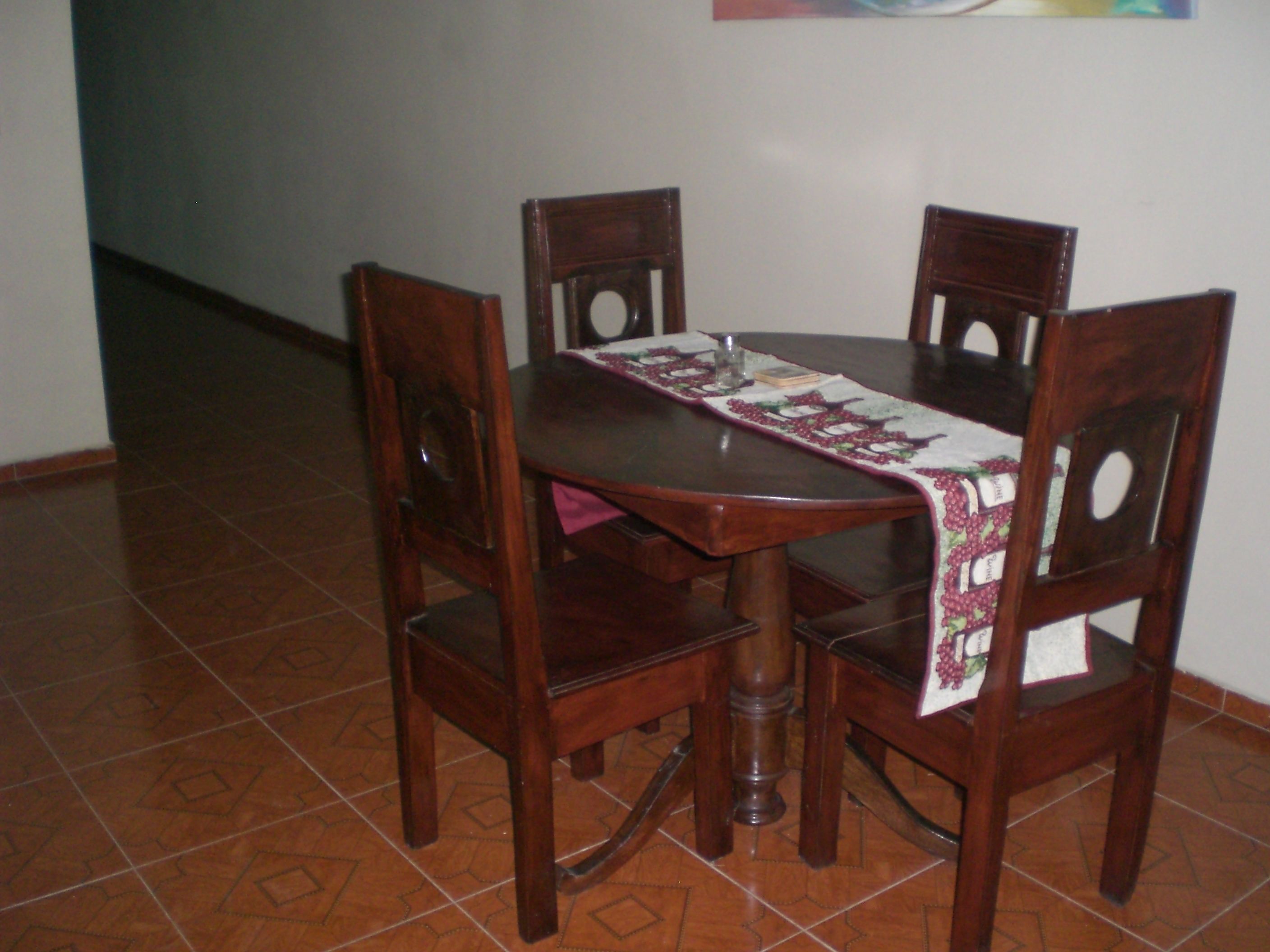 Una mesa de comedor con mantel, cuatro sillas de madera y piso de baldosas.