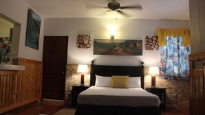 A bedroom with a large bed, two bedside tables with lamps, a ceiling fan, a stone wall, and a painting above the bed. - Metro Residences Hotel (Cap-Haitien)
