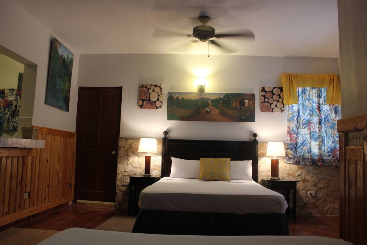 A bedroom with a large bed, two bedside tables with lamps, a ceiling fan, a stone wall, and a painting above the bed.