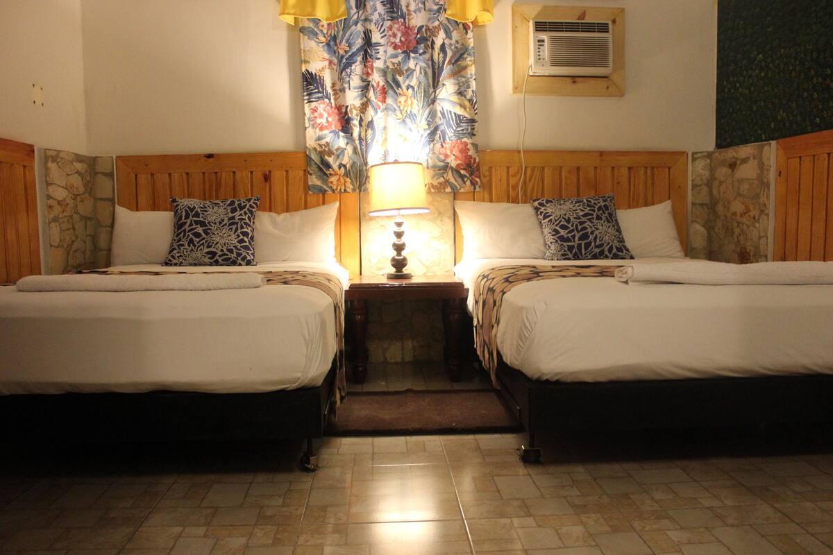 A room with two beds, a nightstand with a lamp, a stone wall, and a floral curtain.