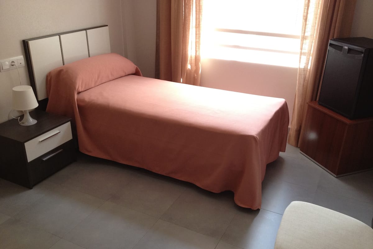A single bed with a pink bedspread, a bedside table with a lamp, a small cabinet, and a television on a stand.