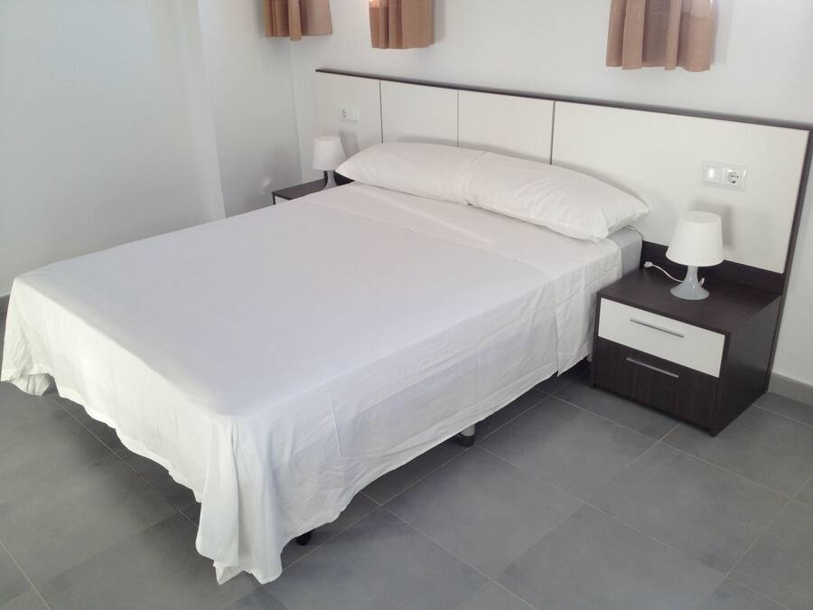 A neatly made bed with white linens, a bedside table with a lamp, and a wall-mounted power outlet.
