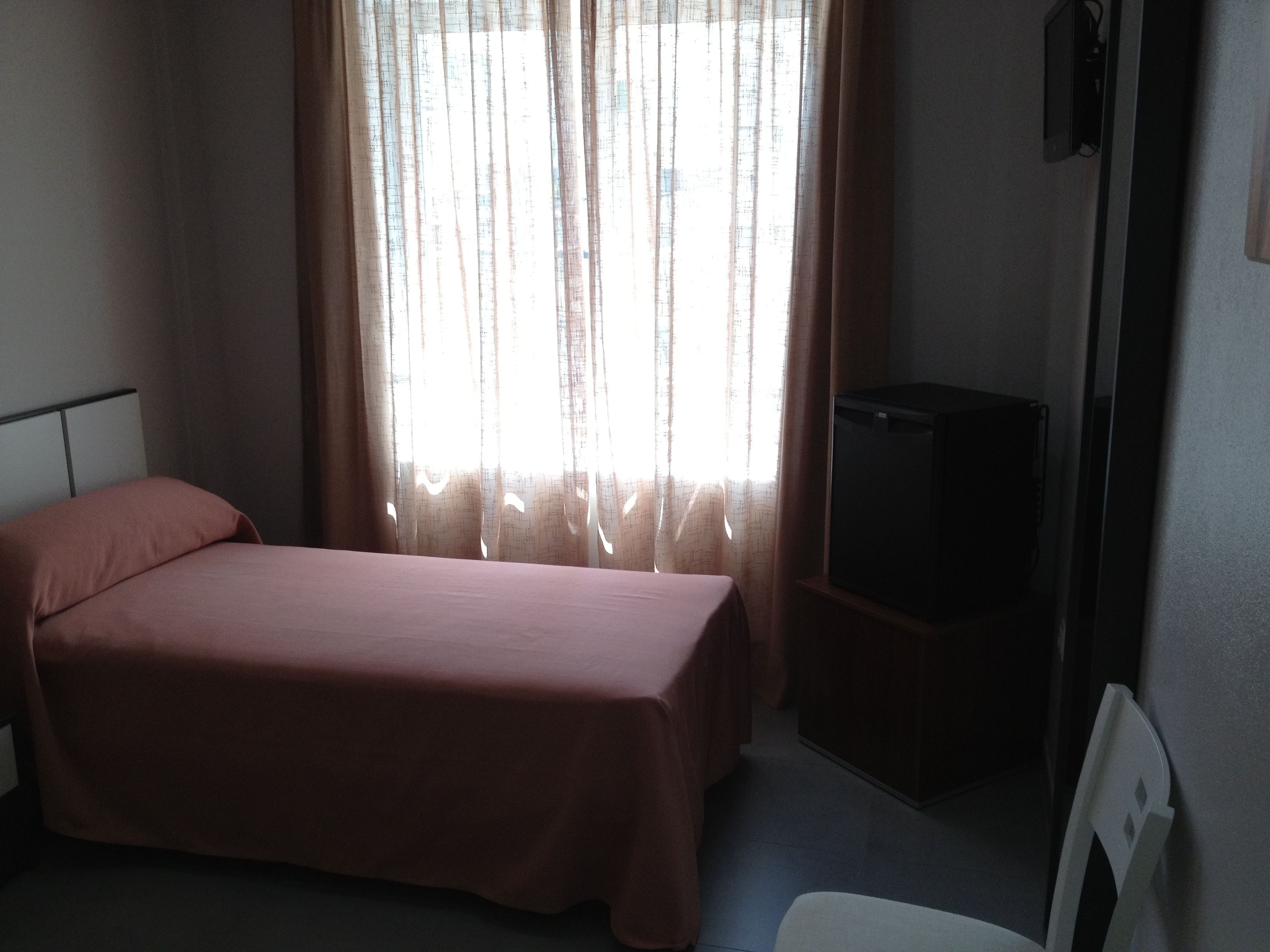 Habitación individual con una cama, un televisor sobre un soporte, una ventana con cortinas translúcidas y una pared lisa.