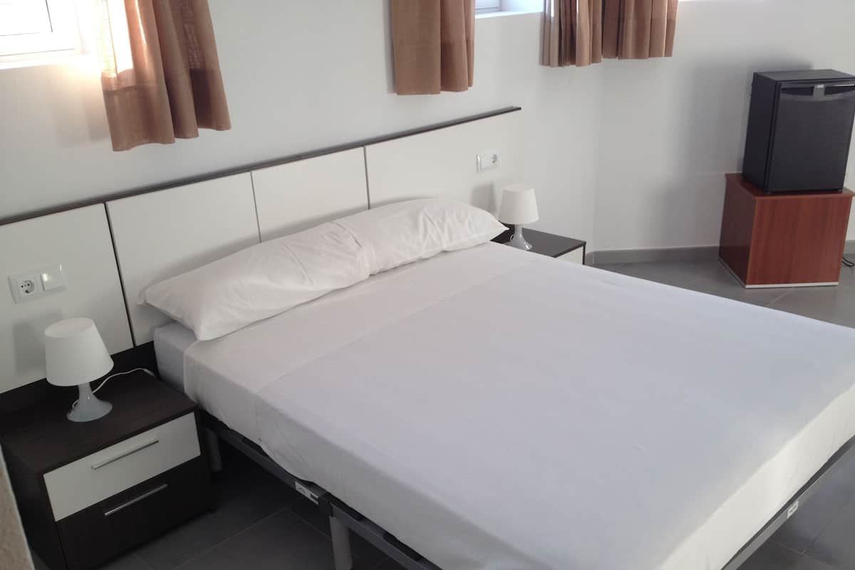 A neatly made bed with white linens, a bedside table with a lamp, a small wooden nightstand, and a television on a stand.