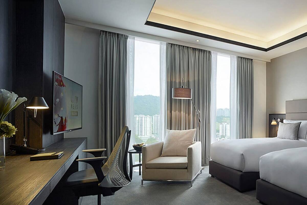 Habitación de hotel con escritorio, silla, cama y ventanas amplias con cortinas.