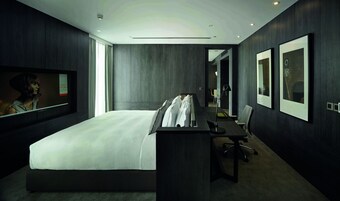 Habitación de hotel moderna con una cama grande, un escritorio y una silla. Hay un televisor en la pared y un cuadro enmarcado.
