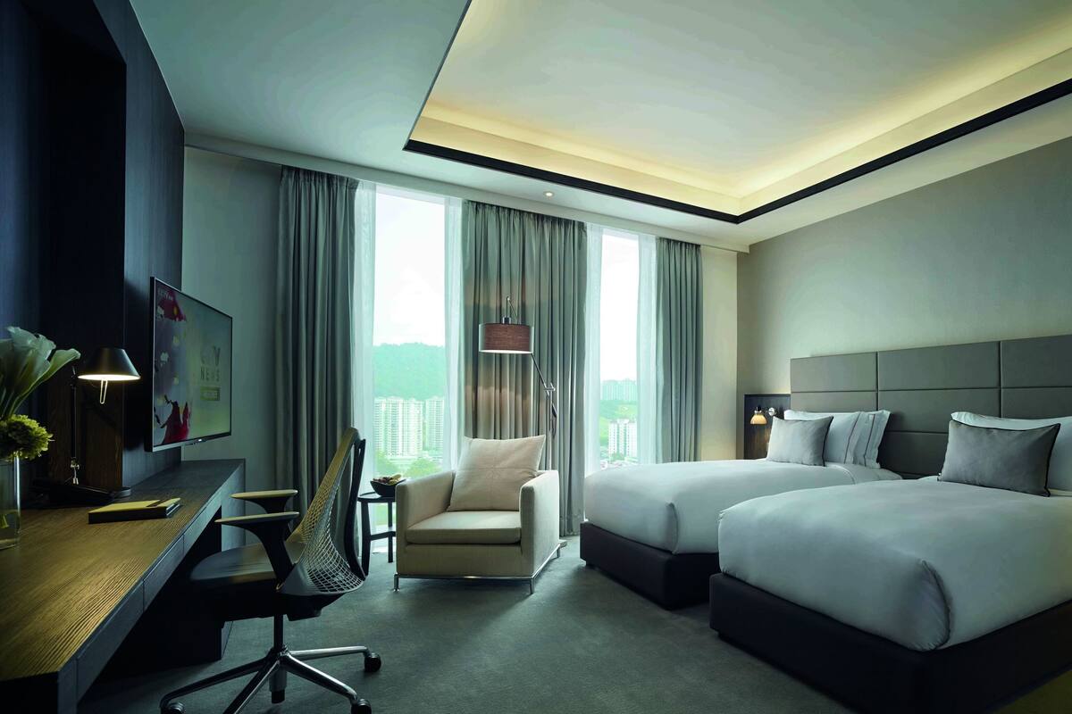 Habitación de hotel con dos camas, un escritorio, una silla, un televisor y vistas a edificios a través de amplias ventanas.