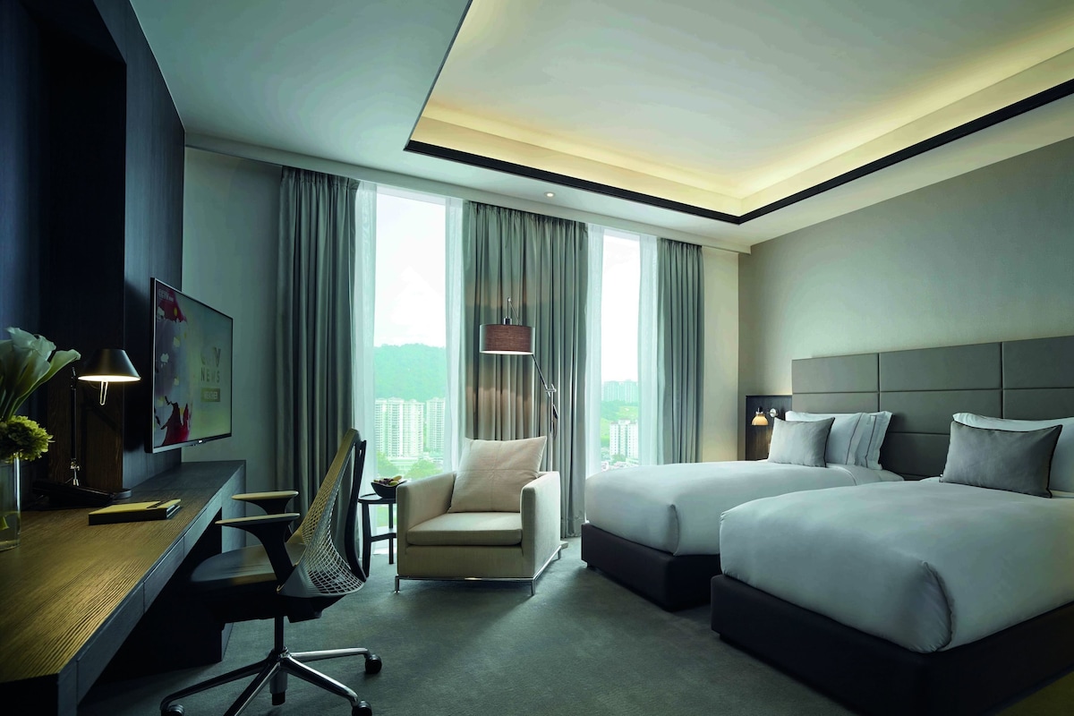 A hotel room with two beds, a desk, a chair, a TV, and a view of buildings through large windows.