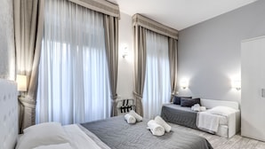 A hotel room with a large bed, a smaller bed, and a desk with a chair. There are curtains on the windows and a lamp on the wall. - Gardenia Suite (Rome)
