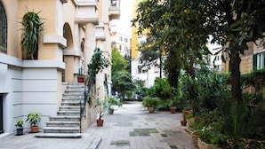 A narrow, paved courtyard with steps leading up to a building entrance, surrounded by potted plants and trees. - Gardenia Suite (Rome)