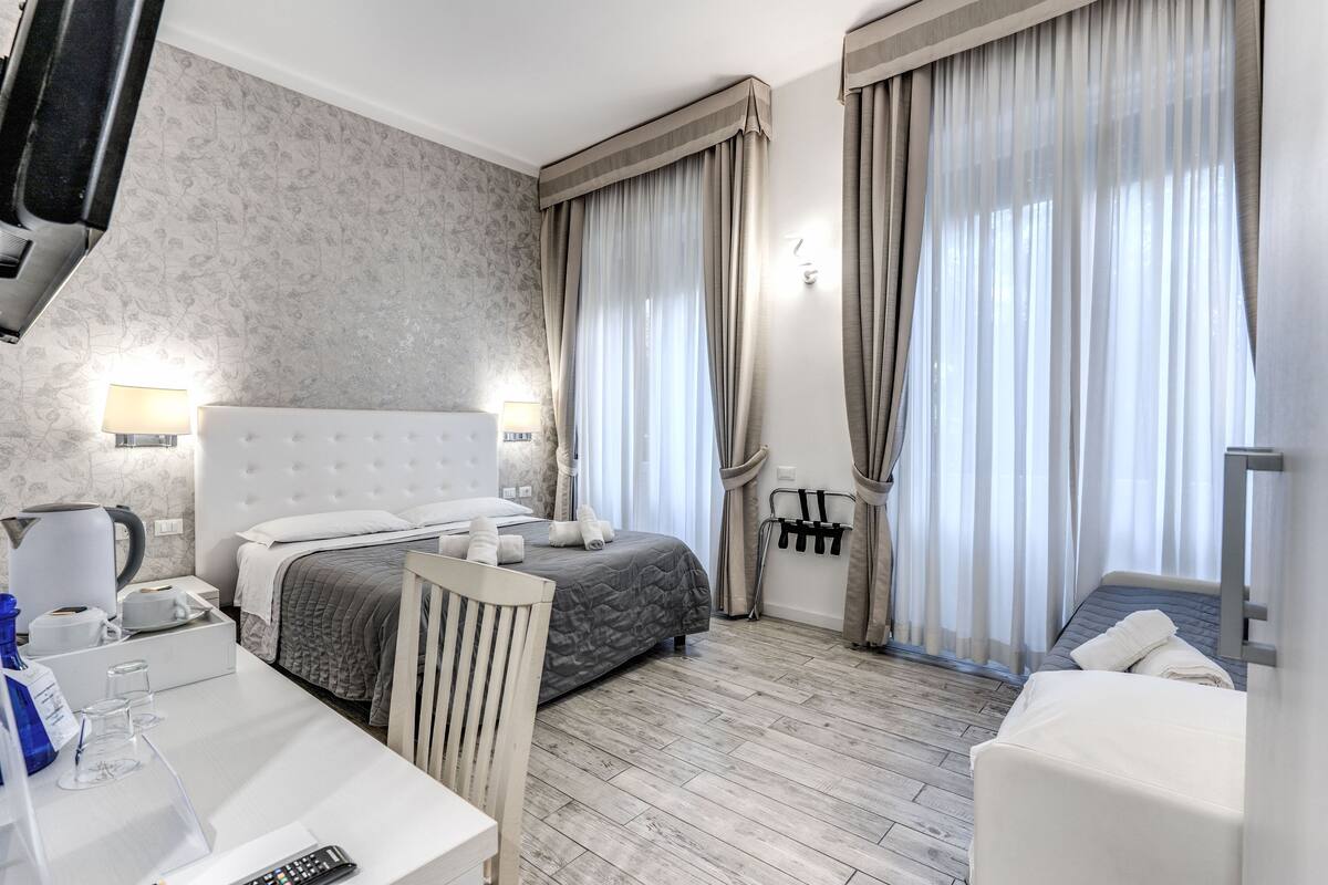 A hotel room with a large bed, a smaller bed, a desk with a lamp, and a chair. There are curtains on the windows and a wall-mounted TV.