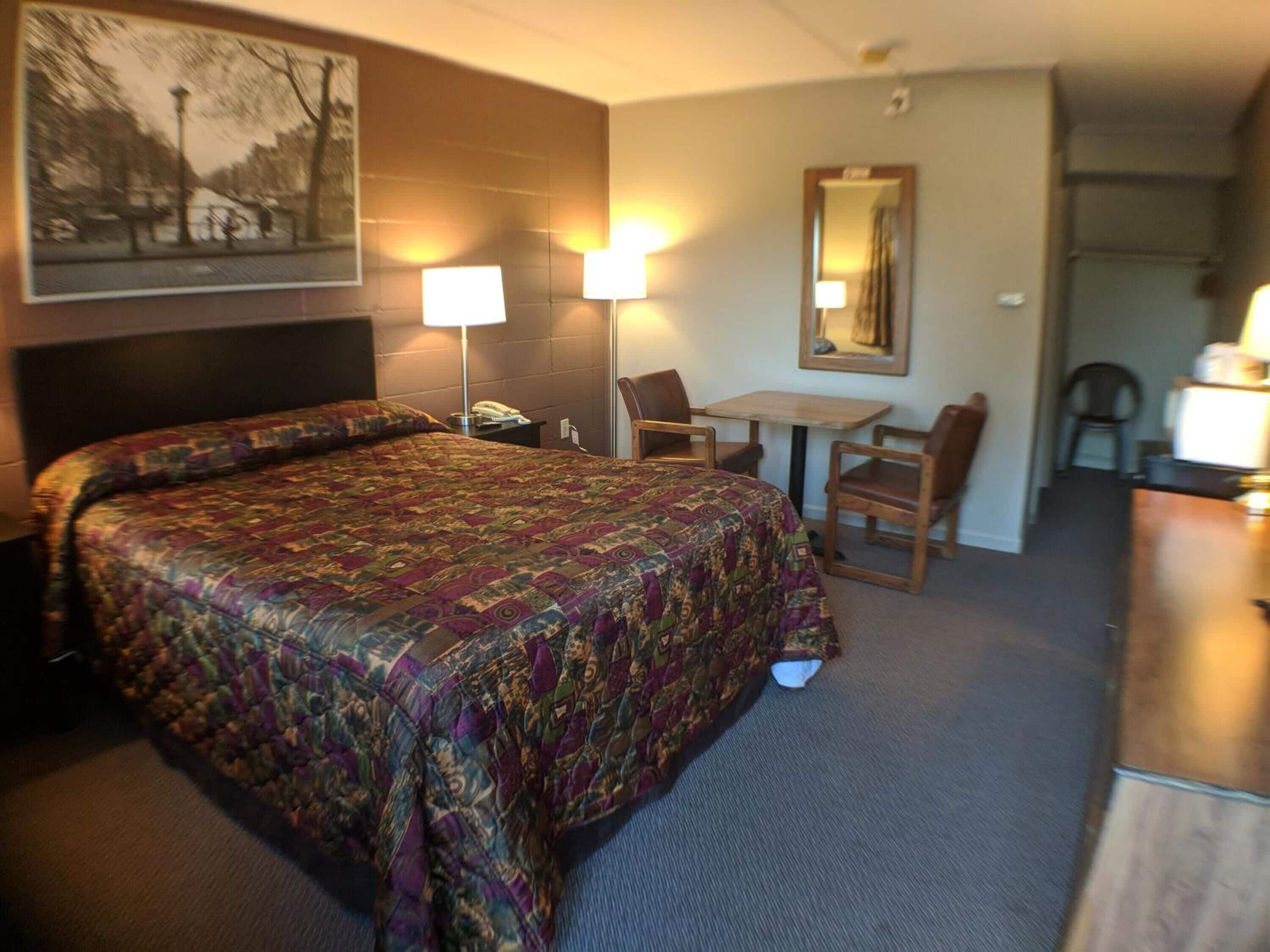 A hotel room with a large bed, a desk, and a chair. There is a lamp on the desk and a mirror on the wall.