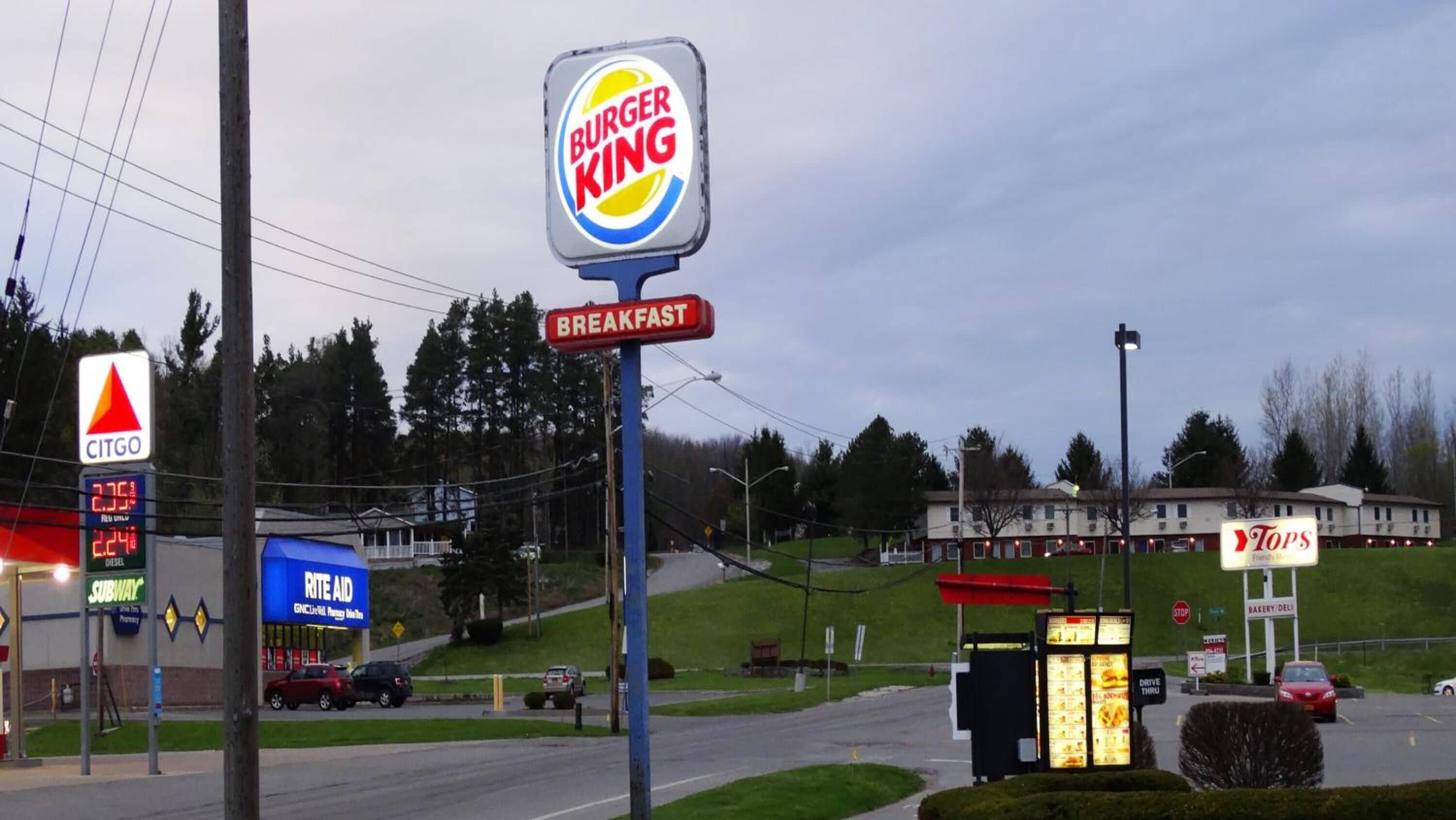 Un cartel de Burger King con un letrero de desayunos debajo, una gasolinera CITGO y una tienda Tops al fondo.