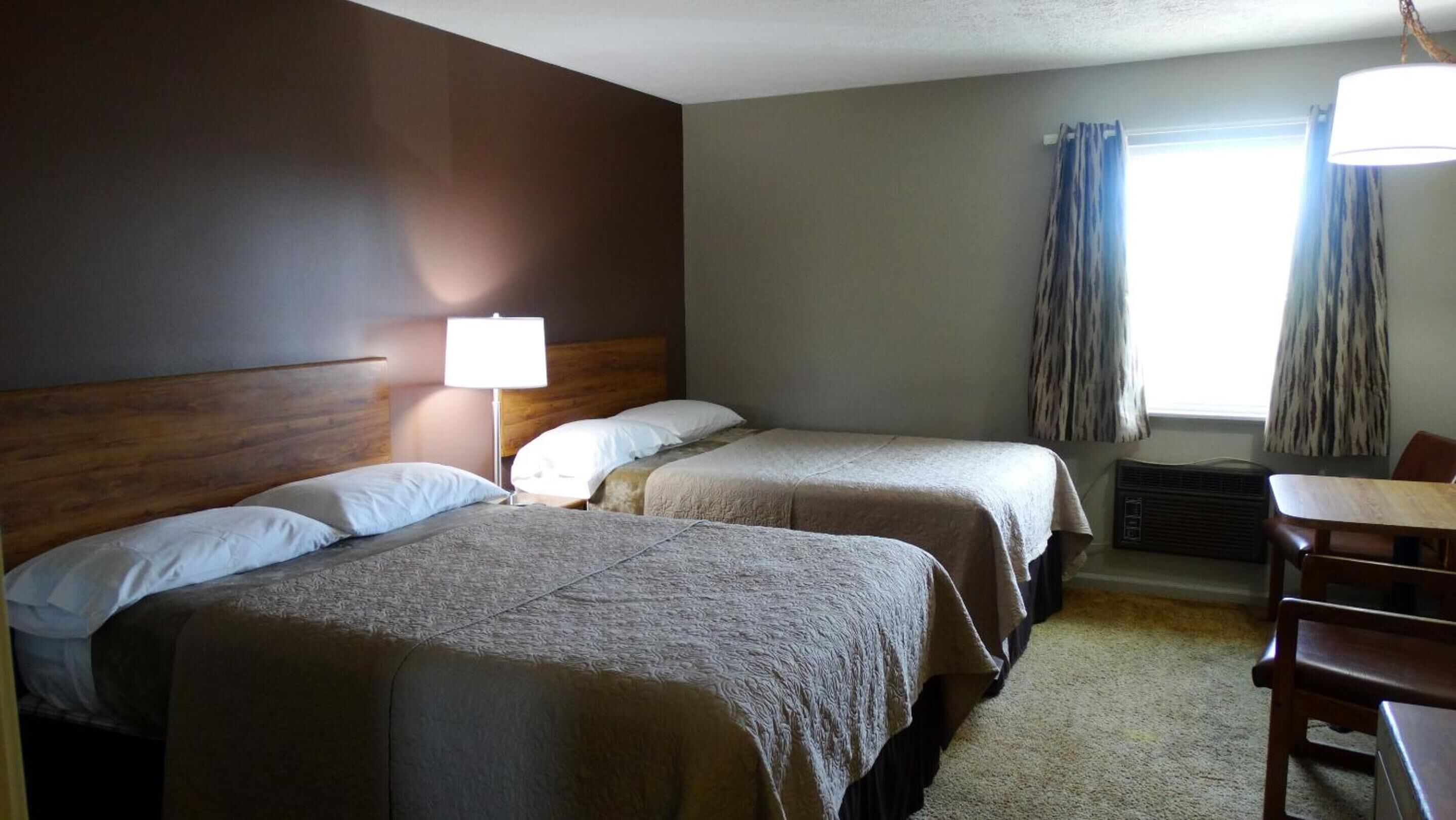 A hotel room with two beds, a desk, a chair, a lamp, and a window with curtains.