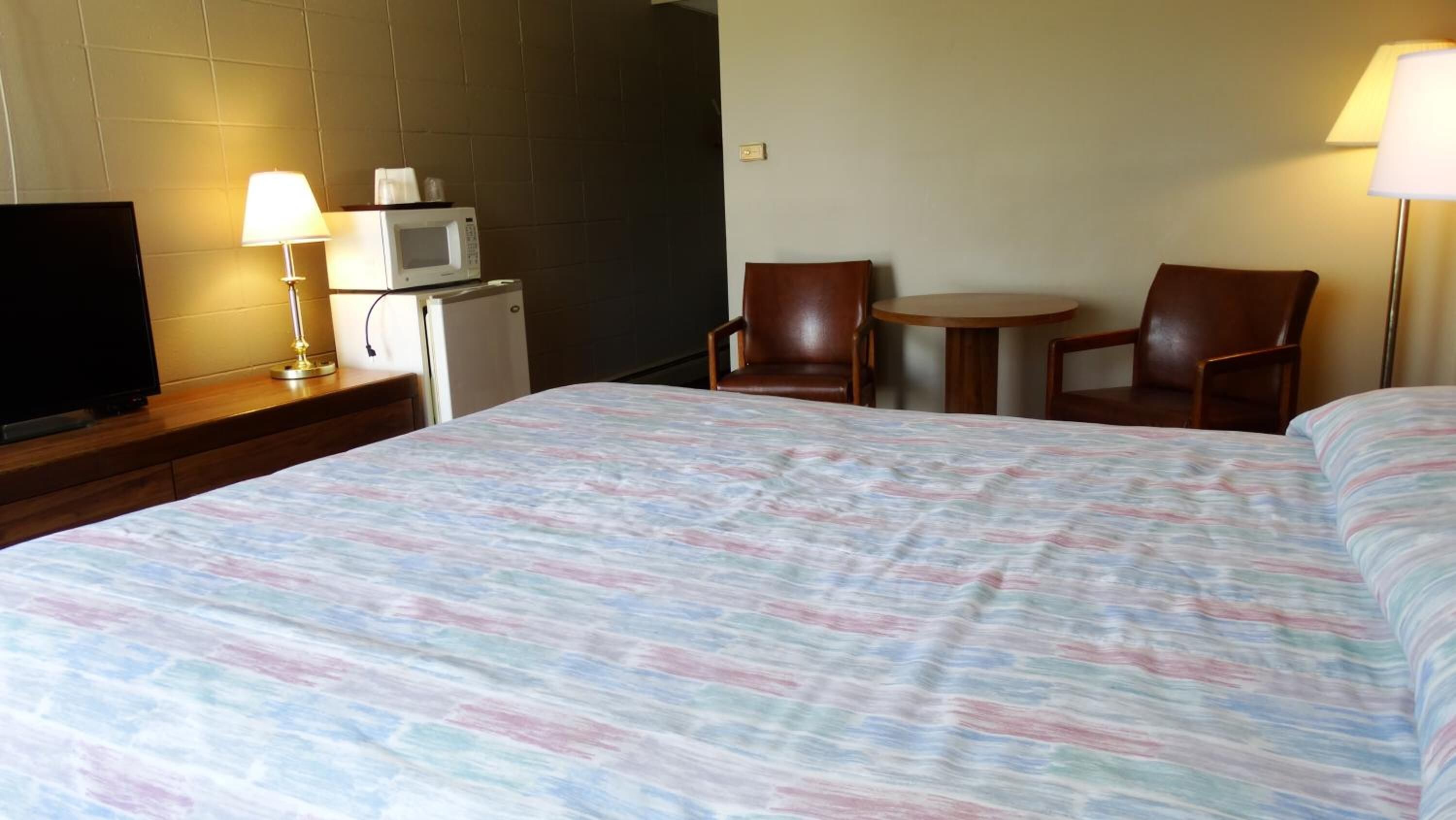 A hotel room with a large bed, a television on a wooden dresser, a microwave, two chairs, and a small table.
