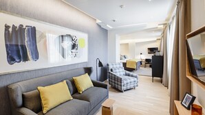 Une chambre d’hôtel moderne avec un canapé gris, un fauteuil à motifs écossais, une table basse en bois et un grand tableau abstrait accroché au mur.