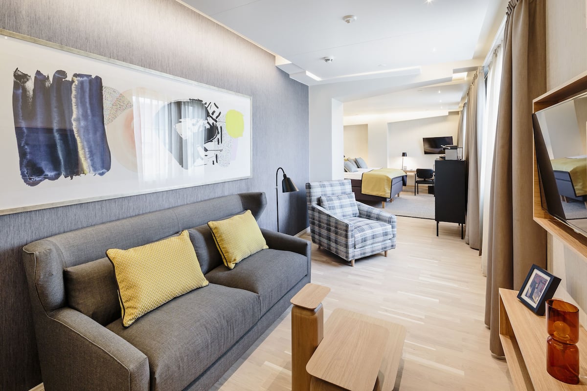 A modern hotel room with a grey sofa, a plaid armchair, a wooden coffee table, and a large abstract painting on the wall.