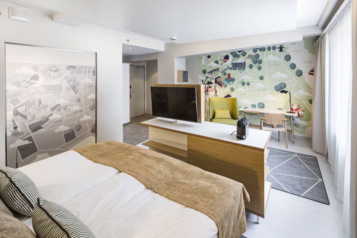 A hotel room with a bed, a desk, a chair, a TV, and a wall with a cityscape mural.