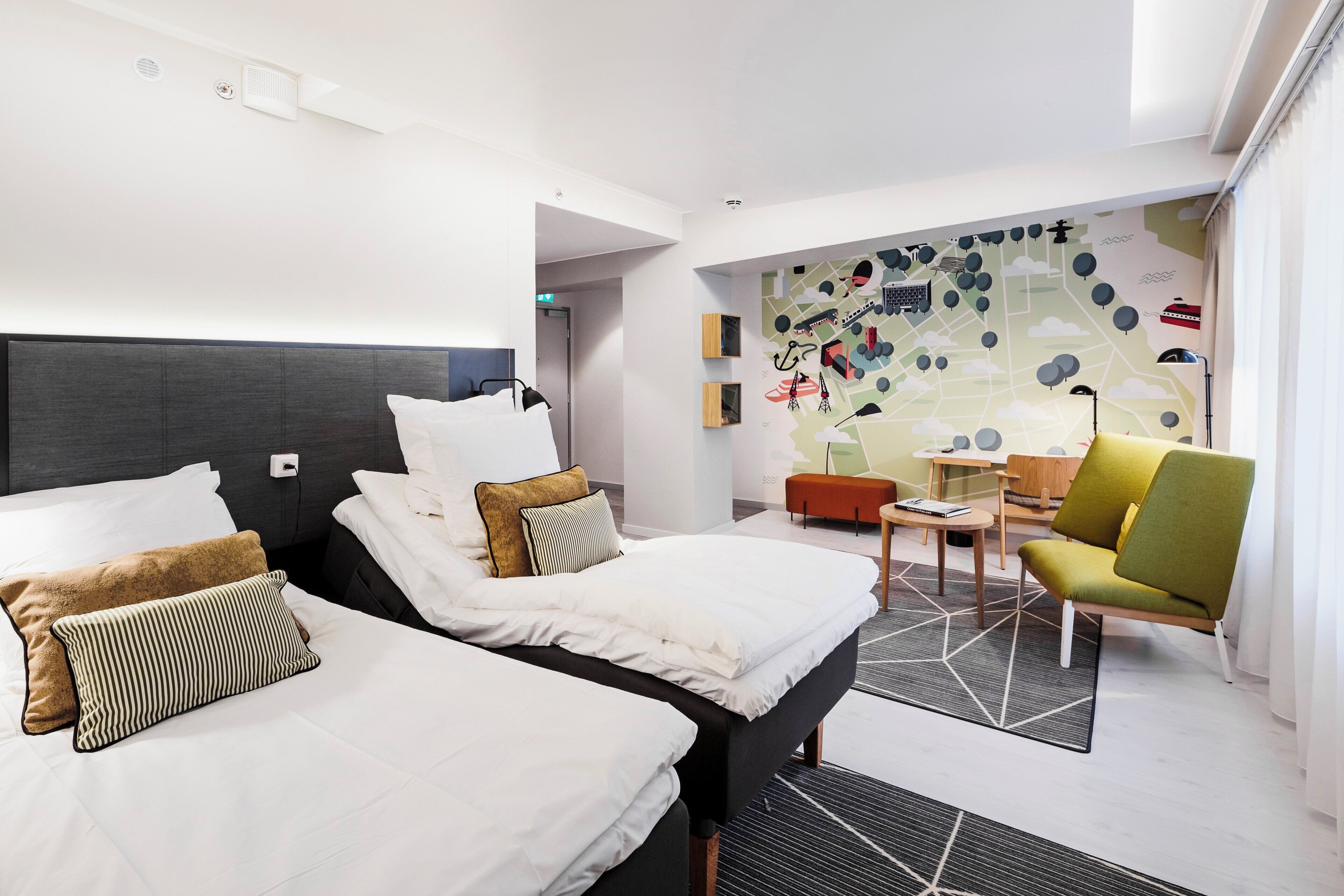 Une chambre d’hôtel moderne avec deux lits, un coin salon comprenant une table et des chaises, et un mur orné d’une fresque représentant un paysage urbain.