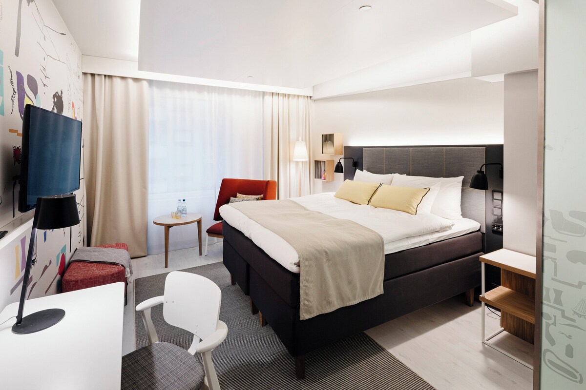 A hotel room with a bed, a desk, a chair, a TV, a lamp, and a window with curtains.
