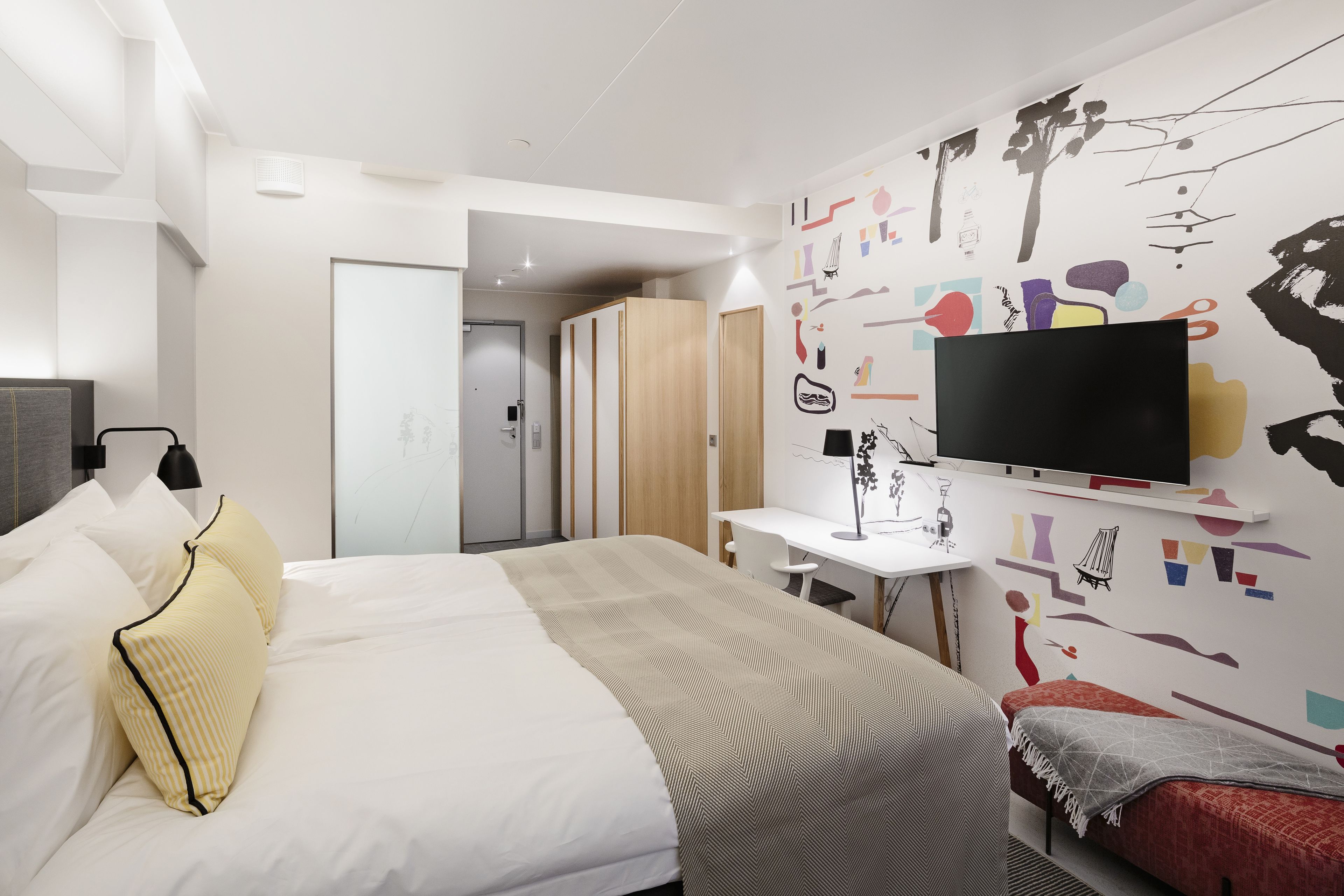 A modern hotel room with a bed, a desk, a television, and a colorful wall mural.