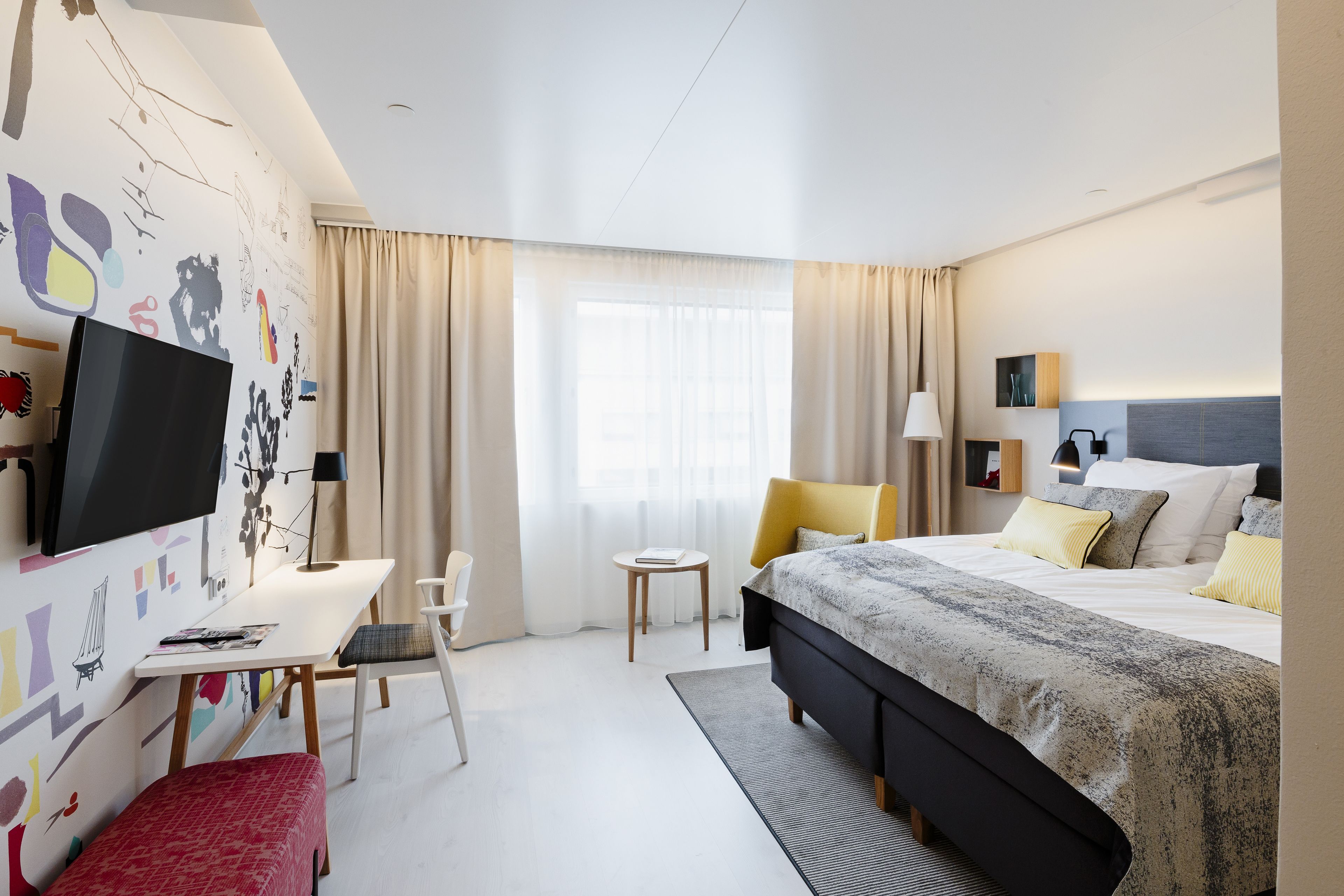 A modern hotel room with a large bed, a desk, a TV, and a window with curtains.