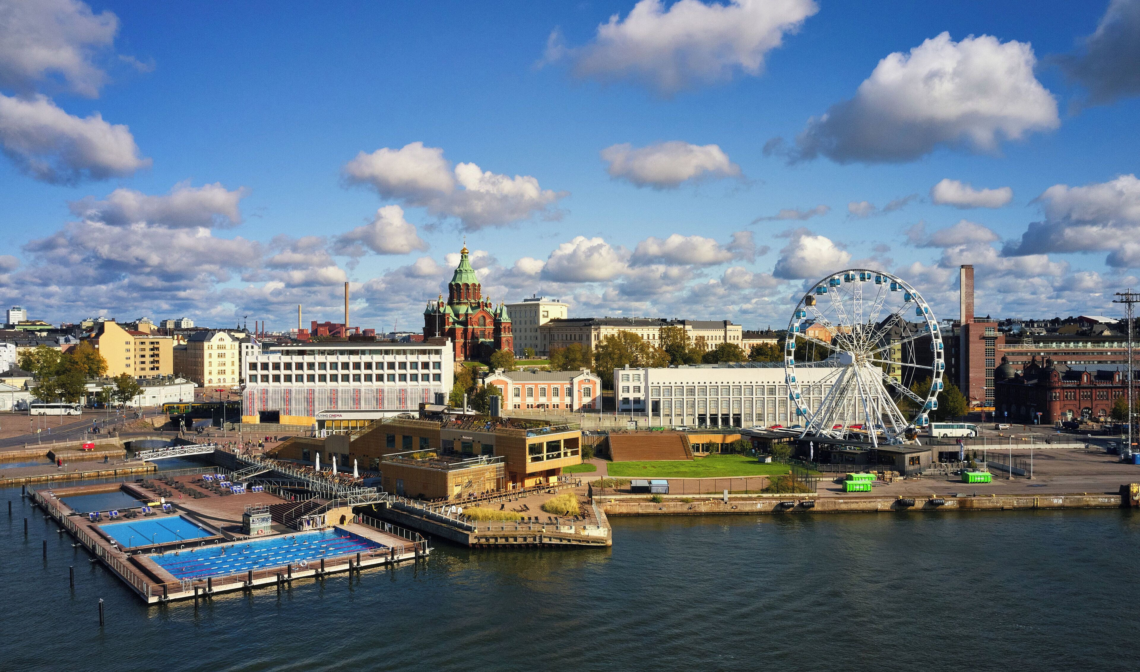 A waterfront cityscape with a prominent Ferris wheel, a swimming pool, and various buildings including a church with a green dome.