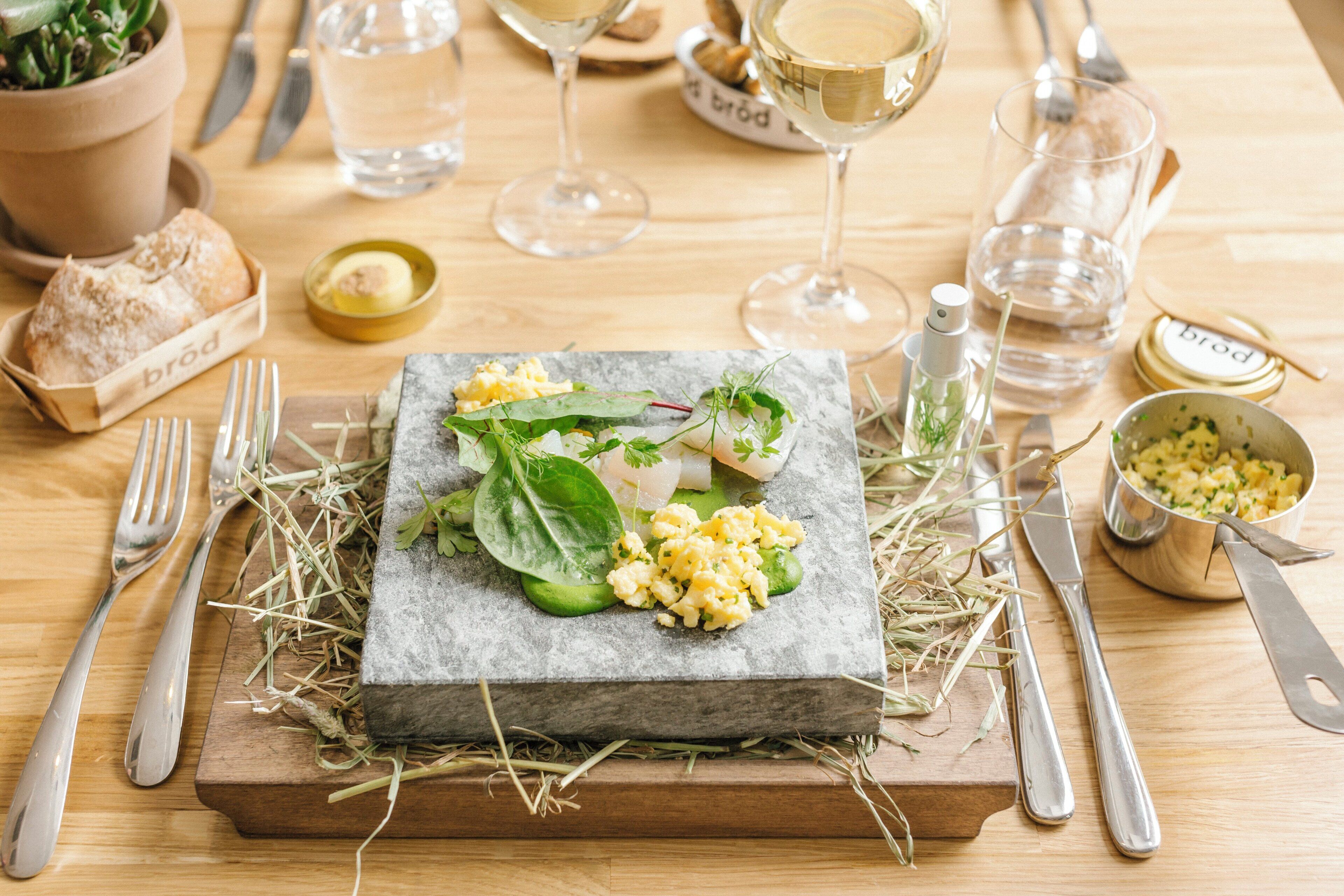 Een houten tafel met een steen dienblad waarop een schotel roerei, groenten en vis liggen, omringd door bestek en een glas witte wijn.