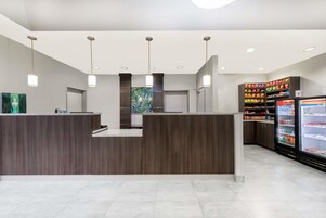Una moderna área de recepción con un mostrador de madera, iluminación colgante y un expositor refrigerado con aperitivos.