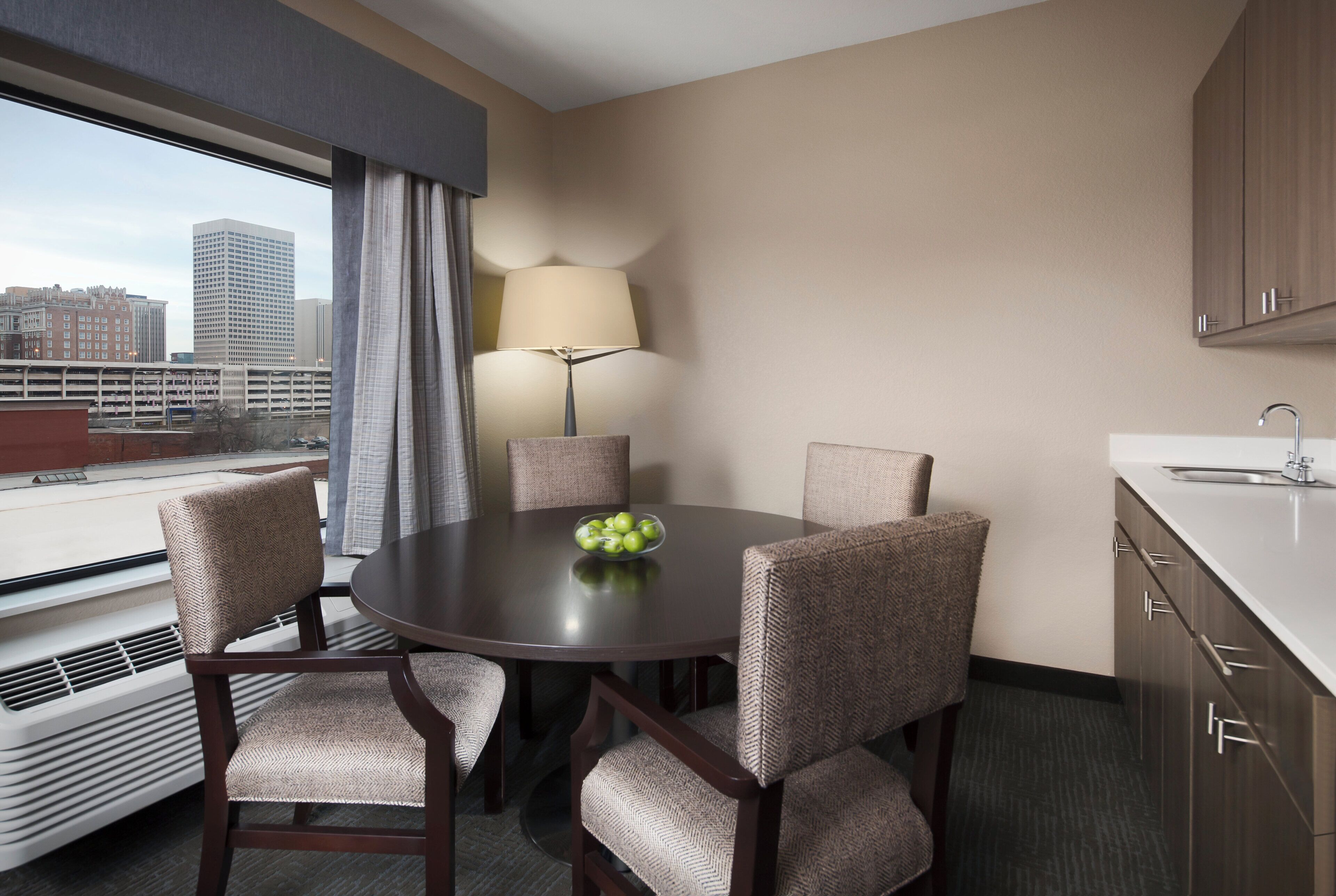 Habitación de hotel con una mesa de comedor, sillas, una lámpara, vistas a edificios y una ventana.