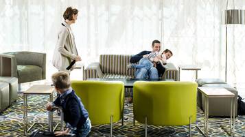 Un vestíbulo de hotel con un sofá a rayas, un sillón verde, un tapete estampado y un niño con una maleta.