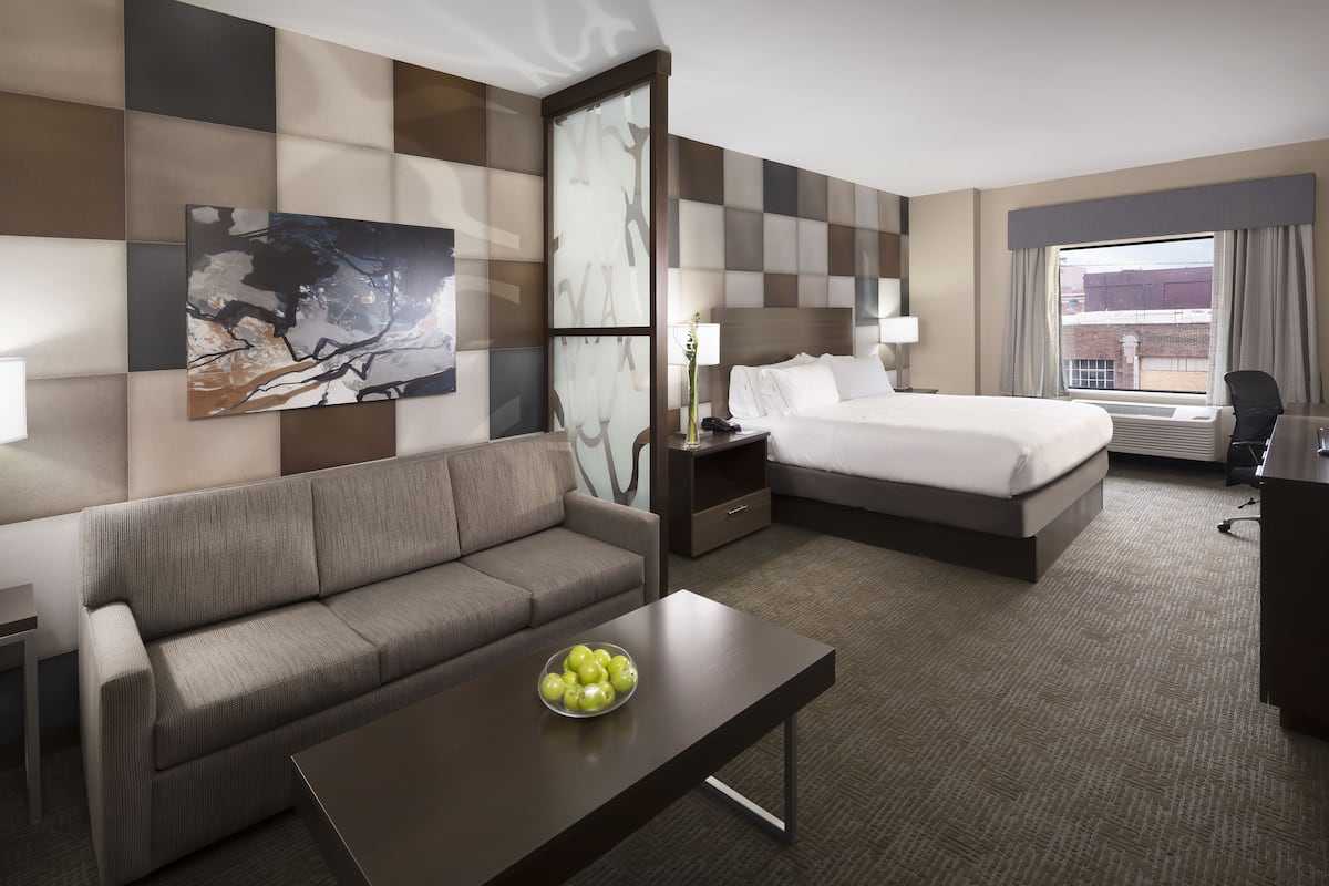 A hotel room with a sofa, a bed, a nightstand, a lamp, and a bowl of fruit on a table.