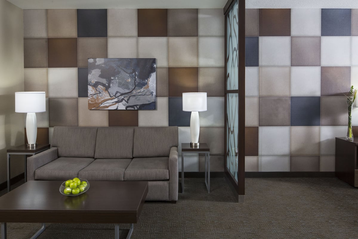A modern hotel room with a grey sofa, two bedside tables with lamps, a coffee table with a bowl of apples, and a large abstract painting on the wall.