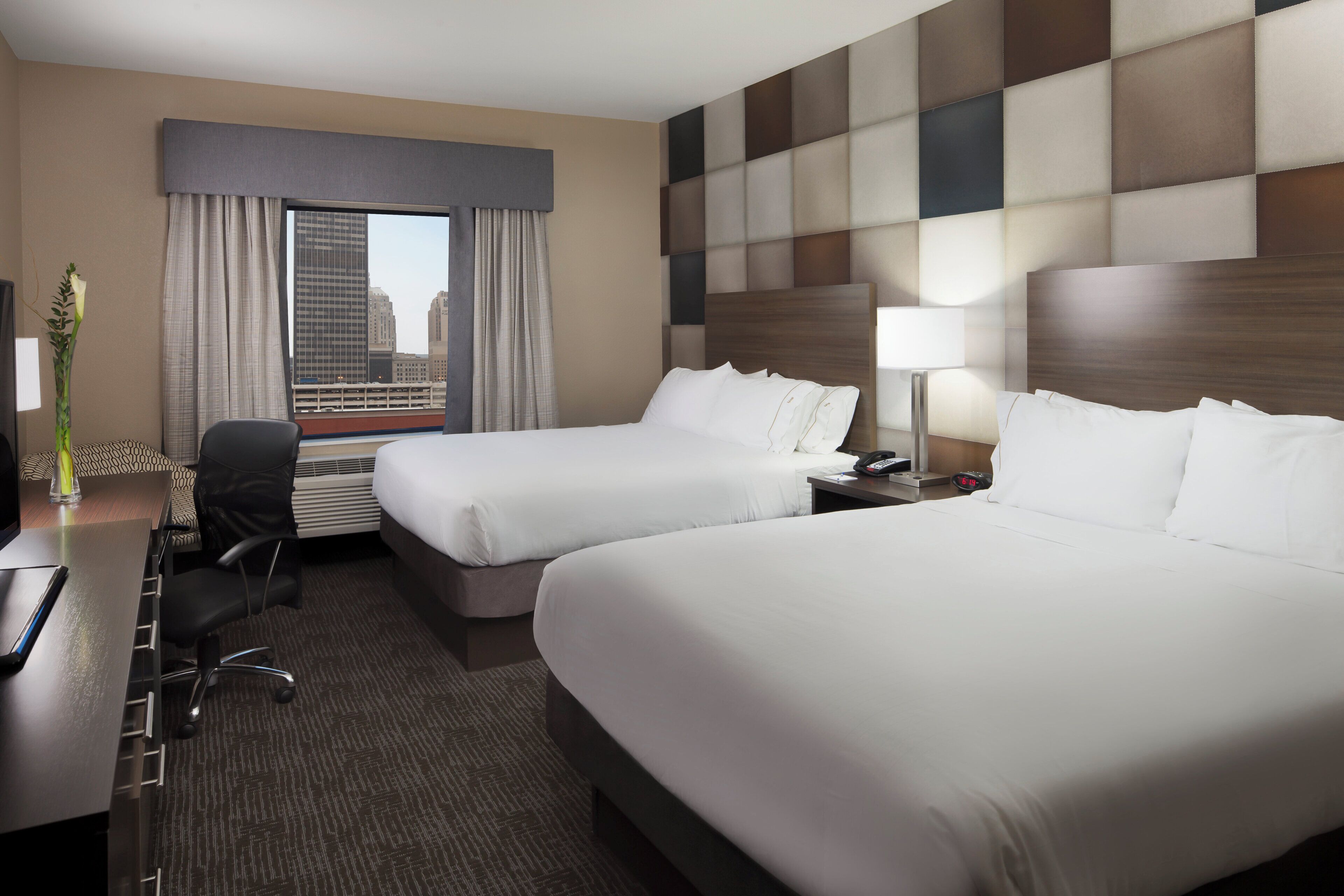 Habitación de hotel con dos camas, un escritorio con silla, una lámpara y vistas a la ciudad.