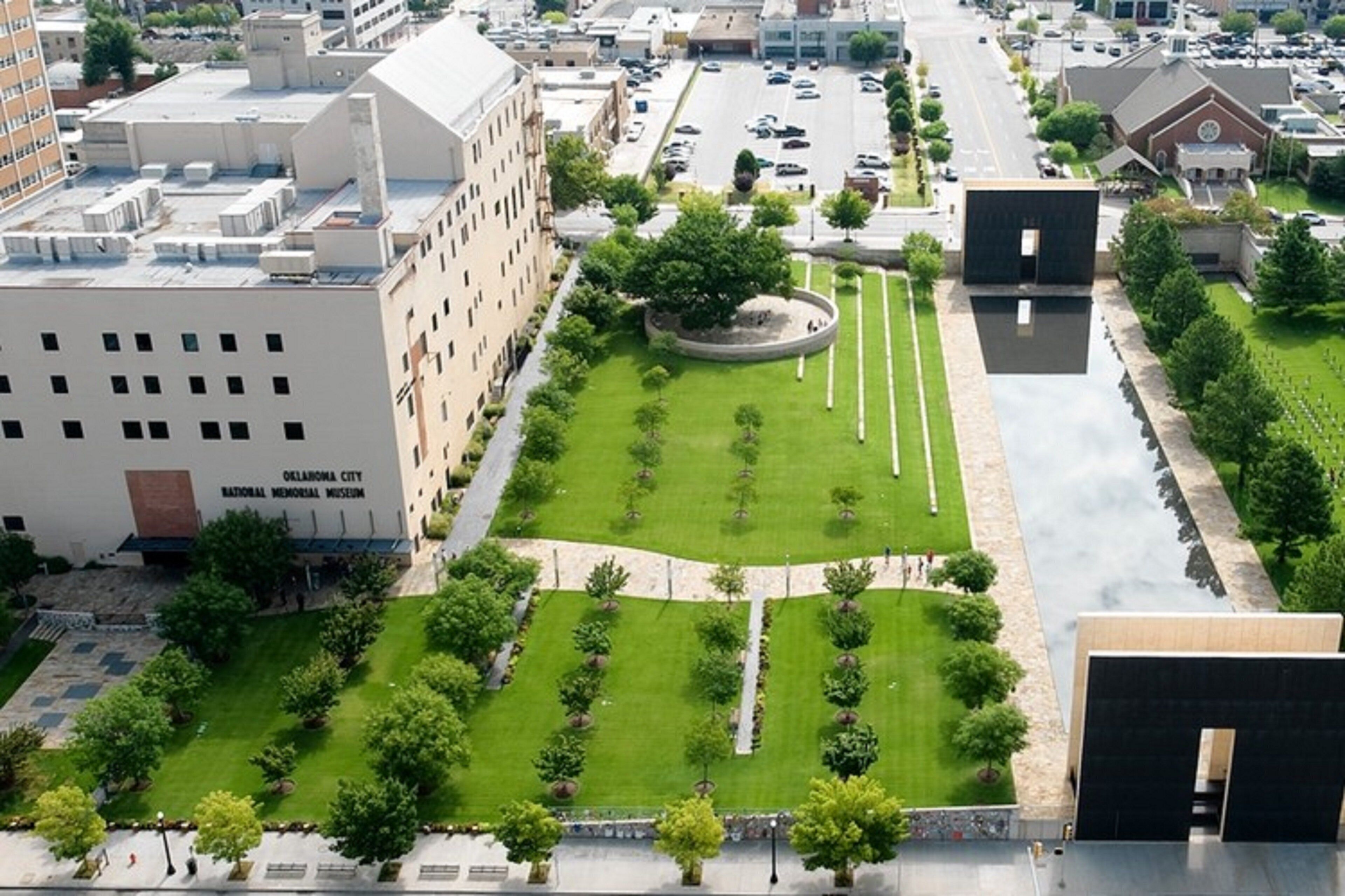 Un área ajardinada con un elemento circular central, rodeada de césped y árboles, adyacente a un edificio con un cartel que dice 'Oklahoma City National Memorial Museum'.