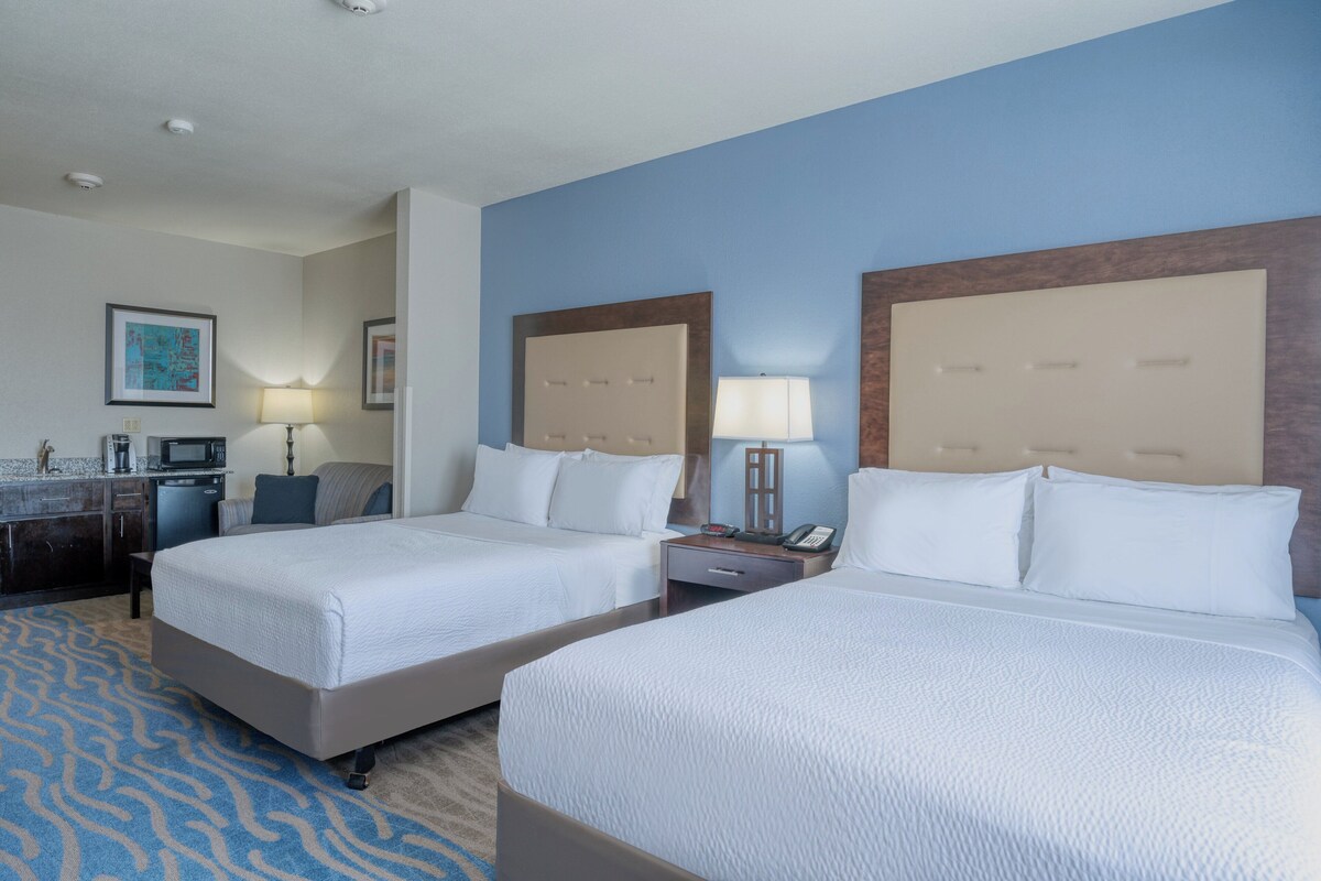 A hotel room with two beds, a desk, a chair, a lamp, and a painting on the wall.