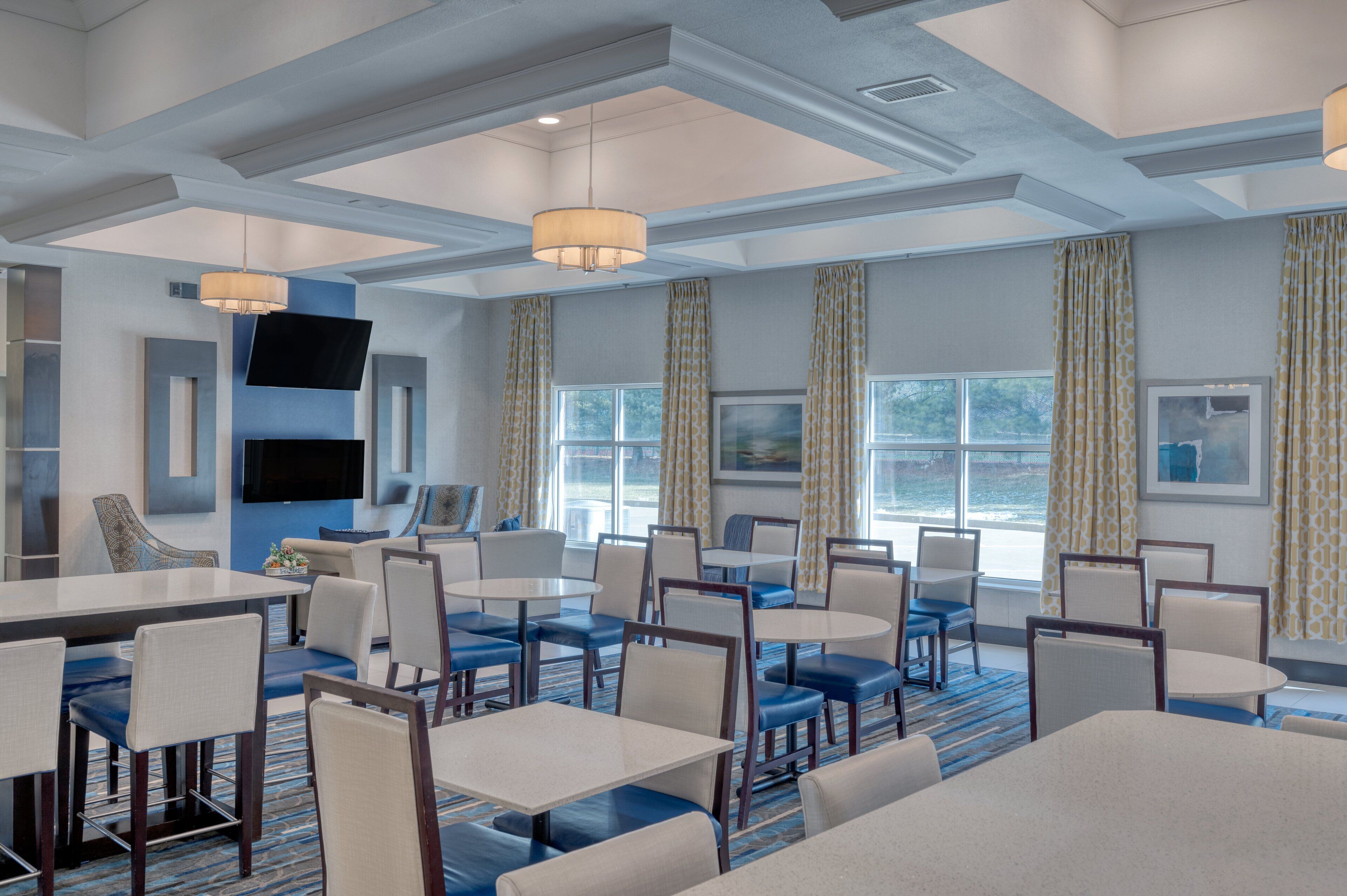 A hotel dining area with multiple tables and chairs, large windows with curtains, and a flat-screen TV mounted on the wall.