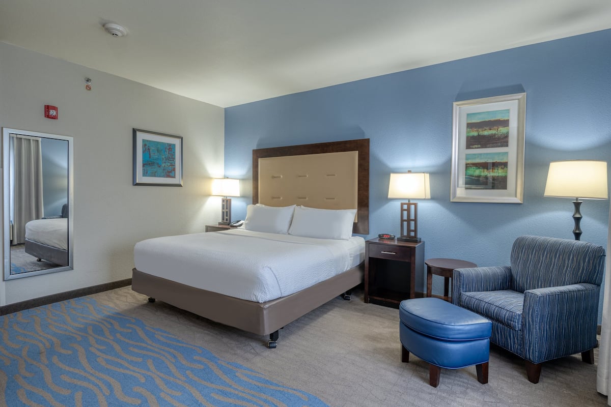 A hotel room with a bed, two bedside lamps, a chair, an ottoman, a mirror, and artwork on the walls.