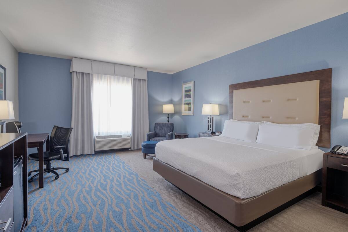 A hotel room with a large bed, a desk with a chair, a small table, and a lamp.