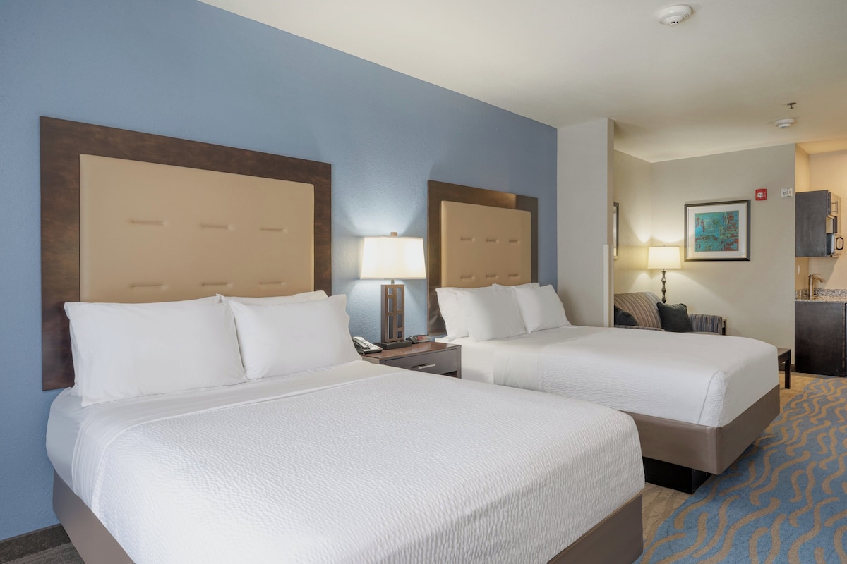 A hotel room with two beds, a nightstand with a lamp, a desk with a chair, a small table, and a painting on the wall.